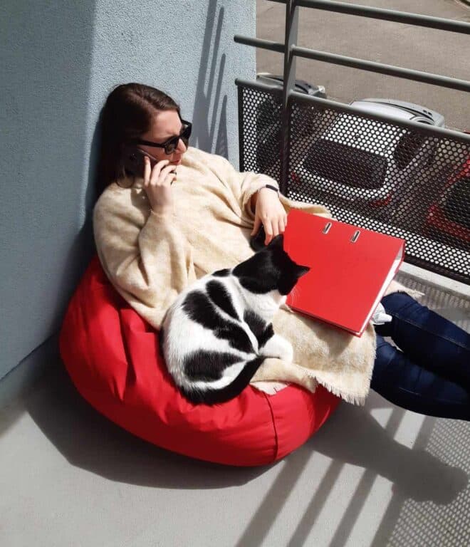 Fille (moi) assise dans un pouf rouge, au téléphone avec un classeur sur les cuisse et un chat noir et blanc (Nessy) couché contre elle.